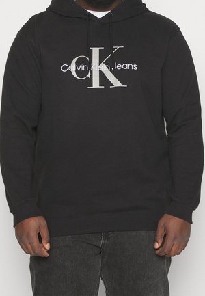 Uomo che indossa una felpa con cappuccio nera con il logo "ck Calvin Klein Jeans" in bianco e grigio chiaro, abbinata a jeans grigio scuro.