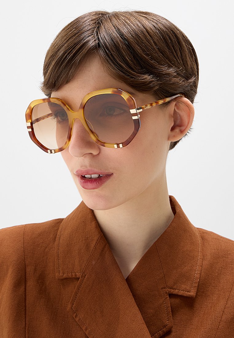 Junge Person mit kurzen braunen Haaren, die übergroße geometrische, bernsteinfarbene Sonnenbrille und eine braune Jacke mit Kragen trägt, schaut leicht nach rechts.