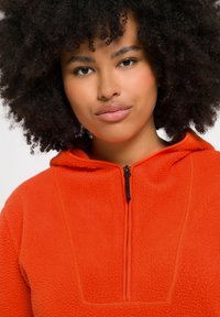 Orange fleece pullover med en lynlåskrave, der har en tekstureret overflade og kontrastsyede detaljer langs fronten og kraven.