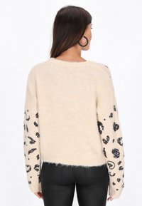 Beżowy sweter dzianinowy z czarnymi, cekinowymi rękawami w leopardzie wzory oraz luźnym krojem. Miękka tekstura, okrągły dekolt, skrócona długość.