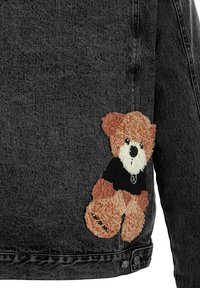 Chaqueta de mezclilla negra con un parche de osito de peluche de pelaje marrón y crema, que lleva puesta una camiseta negra. Costuras detalladas visibles.