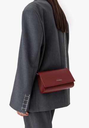 Rote Leder-Umhängetasche mit flachem Design, silberner Kettenriemen und eingraviertem Branding. Kombiniert mit einem grauen, taillierten Blazer mit weißen Nähten.