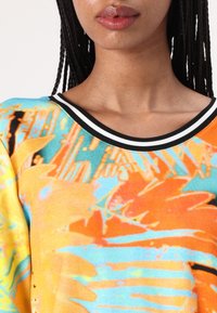 Bunter Pullover in Blau, Orange und Gelb mit abstrakten Mustern. Verfügt über einen schwarz-weißen gestreiften Halsausschnitt. Weicher Stoff.