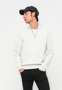 Jeune homme portant une casquette noire, un sweat-shirt blanc à demi-zip et un pantalon noir, se tenant les mains dans les poches, regardant à gauche devant un fond blanc uni.