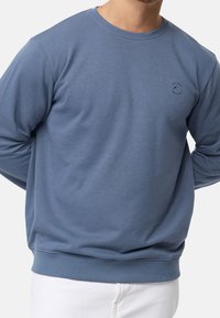Un homme portant un sweatshirt bleu à col rond avec un petit logo sur la poitrine gauche, associé à un pantalon blanc.