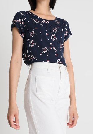 Femme portant un chemisier à manches courtes bleu marine à fleurs, rentré dans un pantalon blanc taille haute avec des poches devant.
