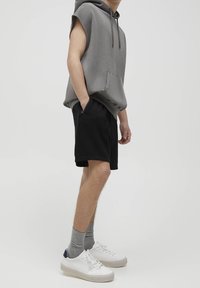 Sweat à capuche sans manches gris avec une poche avant, associé à un short noir. Le mannequin porte des chaussettes grises et des baskets blanches avec des touches de bleu.