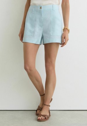 Osoba ubrana w jasnoniebieskie szorty z wysokim stanem i przednimi kieszeniami, brązowe sandały na kostkę z paskiem, biały top oraz złotą bransoletkę.