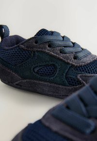 Mörkblå sneakers med nät- och mockamaterial, utrustade med textured detaljer, snören och utskurna designinslag. Närbild.