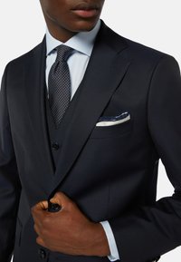 Granatowy garnitur z fakturowanej tkaniny, z białą poszetką z niebieskim obszyciem i krawatem w groszki, uzupełniony jasnoniebieską koszulą.