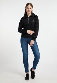 Zwarte sweatshirt met een kwart-rits, geribde kraag en geborduurd ankerlogo, gecombineerd met blauwe skinny jeans en zwarte schoenen.