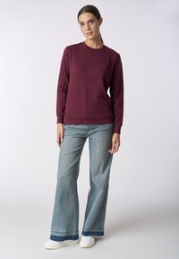 Felpa marrone scuro con maniche lunghe, scollatura rotonda e piccolo emblema. Abbinata a jeans a gamba larga blu chiaro e sneakers bianche.