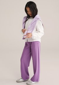 Světle fialový svetr přehozený přes bílou košili s dlouhým rukávem a dekorativním prošíváním. Volné světle fialové kalhoty a stříbrné tenisky.