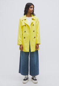 Κίτρινο διπλόπατο αδιάβροχο με μεγάλα κουμπιά, συνδυασμένο με τζιν παντελόνι φαρδιάς γραμμής και μαύρα αθλητικά παπούτσια. Λεία υφή και γυαλιστερό φινίρισμα.