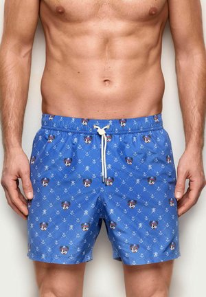 Short de bain bleu avec des visages de dessins animés sur le thème des pirates et des motifs d'ancre blanche porté par un homme torse nu les mains sur les côtés.
