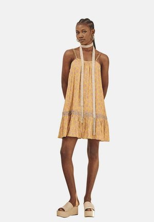 Vestido con estampado floral amarillo, con dobladillo acampanado, tirantes ajustables y lazos decorativos. La modelo lleva sandalias de plataforma.