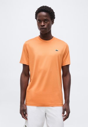 Muž oblečený v priliehavom oranžovom tričku s krátkym rukávom so zeleným malým logom krokodíla a bielych šortkách, stojaci pred jednoduchým pozadím.