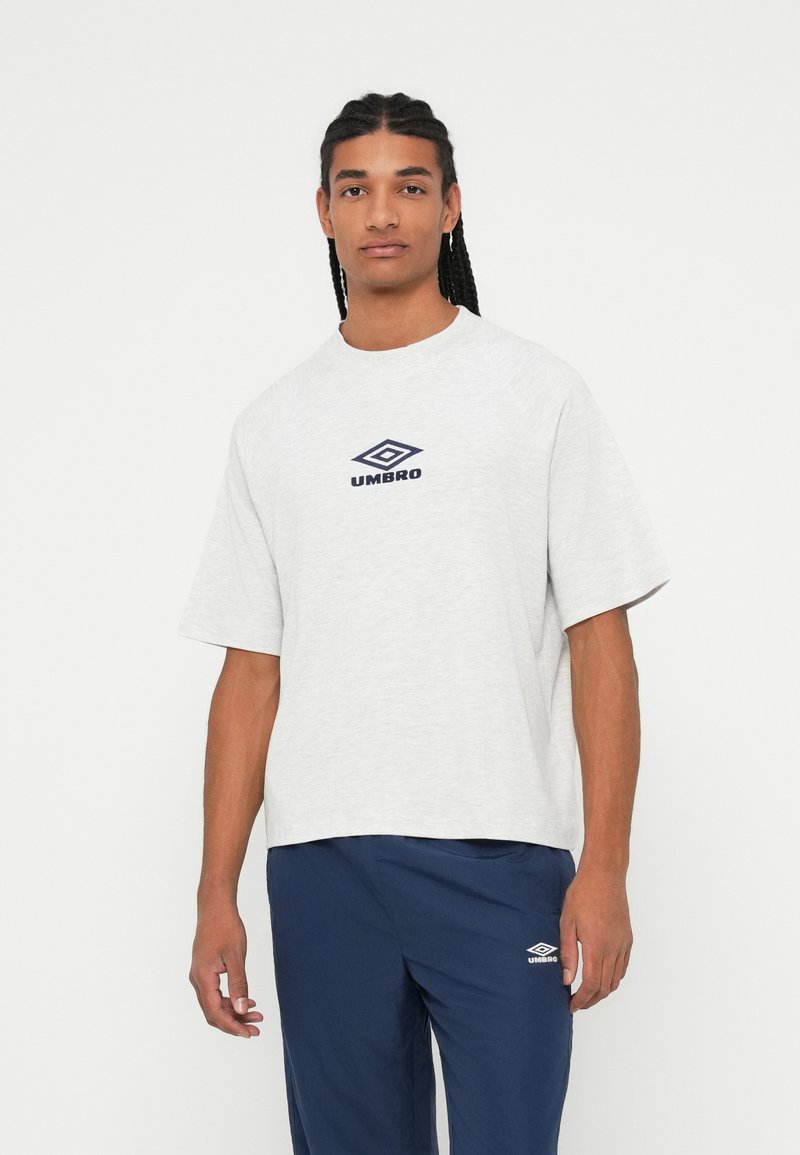 Giovane uomo con capelli intrecciati che indossa una maglietta grigia chiaro Umbro e pantaloni blu navy Umbro, in piedi su uno sfondo bianco.