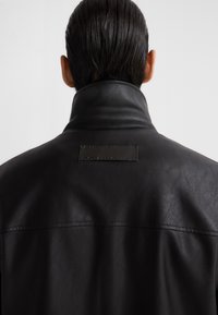 Giacca di pelle nera con colletto alto, texture liscia e un'etichetta rettangolare con il nome "ADULFO DOMINGUEZ" sul retro.