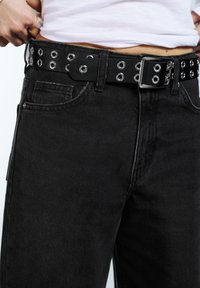 Čierne denimové džínsy s textúrovaným povrchom, vybavené čiernym opaskom zdobeným striebornými okami a kovovou prackou, detailný pohľad.