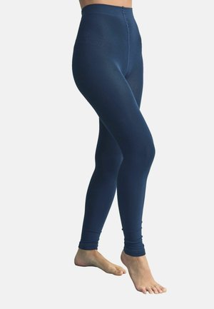 Osoba boso, ubrana w granatowe legginsy z wysokim stanem, stojąca z jedną stopą lekko uniesioną, na jednolitym jasnym tle.