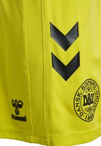 Gul sportströja gjord av slätt tyg, med svarta chevronmönster och en cirkulär logotyp med text. Varumärket "Hummel" längst ner.