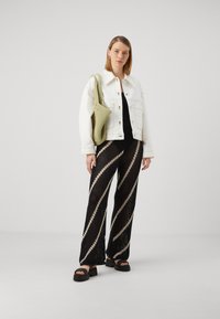 Veste en jean blanche sur un haut noir, associée à un pantalon en crochet rayé noir et beige et des sandales noires épaisses. Sac tissé vert en accessoire.