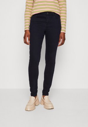 Personne portant un pantalon slim noir, des mocassins chunky beiges et une chemise à manches longues rayée verte et rose, se tenant devant un fond blanc.