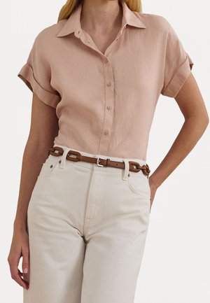 Kobieta nosząca jasnoróżową koszulę z krótkim rękawem, wpuszczoną w białe spodnie z wysokim stanem i brązowym paskiem.