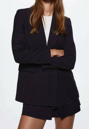 Nainen, joka ristii kädet, pukeutuneena mustaan raitakuviolliseen bleiseriin ja siihen sopiviin shortseihin valkoisen paidan päällä, seisoo yksinkertaista taustaa vasten.