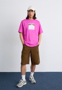 Lys pink T-shirt i bomuld med grafisk print, parret med brune cargo shorts, hvide ankelsokker og grå sneakers. Lys kasket på hovedet.
