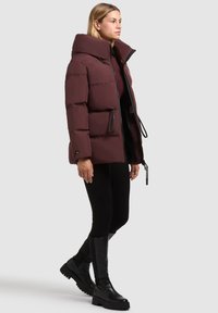 Chaqueta de plumas burdeos con cuello alto, acentos negros, cierre de cremallera y dos bolsillos laterales. Textura suave y forma cuadrada con cintura ajustada.