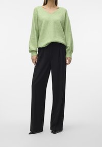 Hellgrüner Oversize-Pullover mit V-Ausschnitt, kombiniert mit weit geschnittenen schwarzen Hosen. Stricktextur, lange Ärmel und spitze schwarze Schuhe.