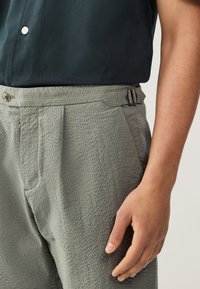 Pantaloni verdi leggeri e testurizzati con pieghe; caratterizzati da un bottone e fibbie regolabili in vita.