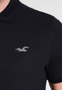 Černé polo tričko s texturovanou tkaninou, ohnutým límečkem a malým šedým vyšitým logem ptáka na levé straně hrudi.