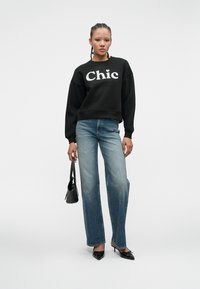 Woman stands wearing black "Chic" sweatshirt, faded straight-leg jeans, black pointed heels, and holding a small black handbag against white background.