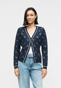 Ναυτικό ζακέτα με λευκές λεπτομέρειες, με κεντημένα σχέδια. Κατασκευασμένη από απαλό υλικό, διαθέτει V-λαιμόκοψη και κλείσιμο με κουμπιά, φορεμένη πάνω από ένα λευκό πουκάμισο.