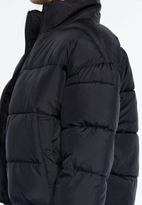 Zwarte pufferjas met hoge kraag, gequiltde textuur en elastische manchet. Bevat een ritssluiting en zijzakken voor functionaliteit.