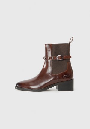 Bottine en cuir marron avec panneaux latéraux élastiques, bout carré, boucle décorative avec clous et un talon épais. Texture lisse et design minimal.
