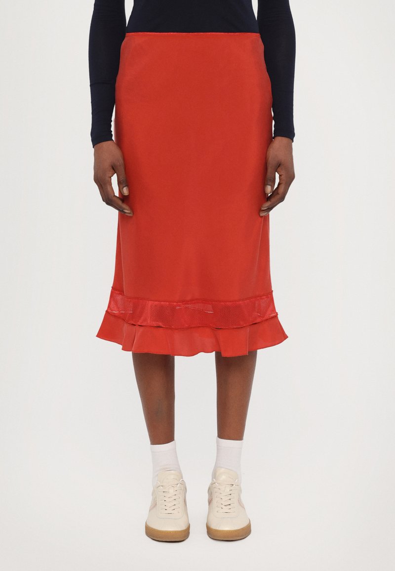 Red midi skirt with a sheer ruffled hem, smooth texture, and fitted silhouette. Paired with white sneakers and ankle-length socks.