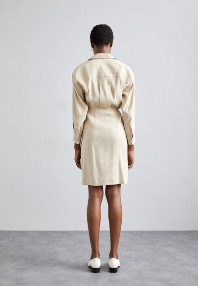 Vestido beige de botones con mangas largas, cintura ceñida y dobladillo a la altura de la rodilla. La modelo lleva zapatos blancos, vista desde la parte de atrás.