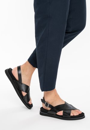 Füße mit schwarzen Leder-Sandalen mit überkreuzten Riemen und verstellbarer Schnalle, kombiniert mit dunkelblauen strukturierten Hosen vor weißem Hintergrund.