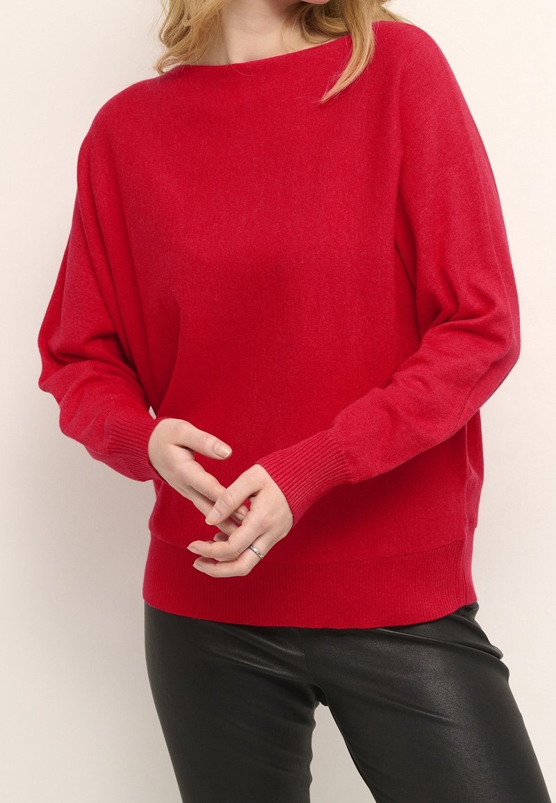 Pull en maille rouge avec un col rond, manches longues, et poignets et ourlet côtelés. Porté avec un pantalon en similicuir noir, texture douce visible.