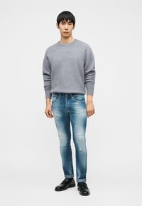 Άνδρας στέκεται φορώντας ένα ανοιχτό γκρι μάλλινο πουλόβερ, ξεθωριασμένο στενό μπλε τζιν και μαύρα δερμάτινα μοκασίνια σε λευκό φόντο.