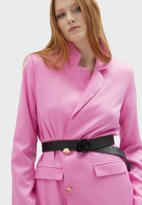 Rosa kavaj med en strukturerad krage och dubbelknäppt design, pryd med guldfärgade knappar. Kombinerad med ett svart texturerat bälte och fickor.