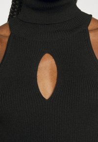 Gros plan d'une personne portant un haut noir côtelé sans manches à col roulé avec une découpe en forme de trou à la poitrine.