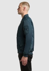 Ung man i blå bomberjacka och svarta byxor står i profil mot en enkel ljusgrå bakgrund.