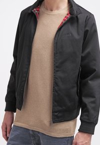 Svart bomberjacka med röd rutigt foder, dragkedja framtill, ribbade ärmslut och nederkant, tillsammans med en beige strukturerad tröja under.