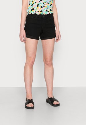 Mujer con pantalones cortos negros con dobladillo enrollado, una camiseta de patrón abstracto multicolor y sandalias negras, de pie frente a un fondo liso.