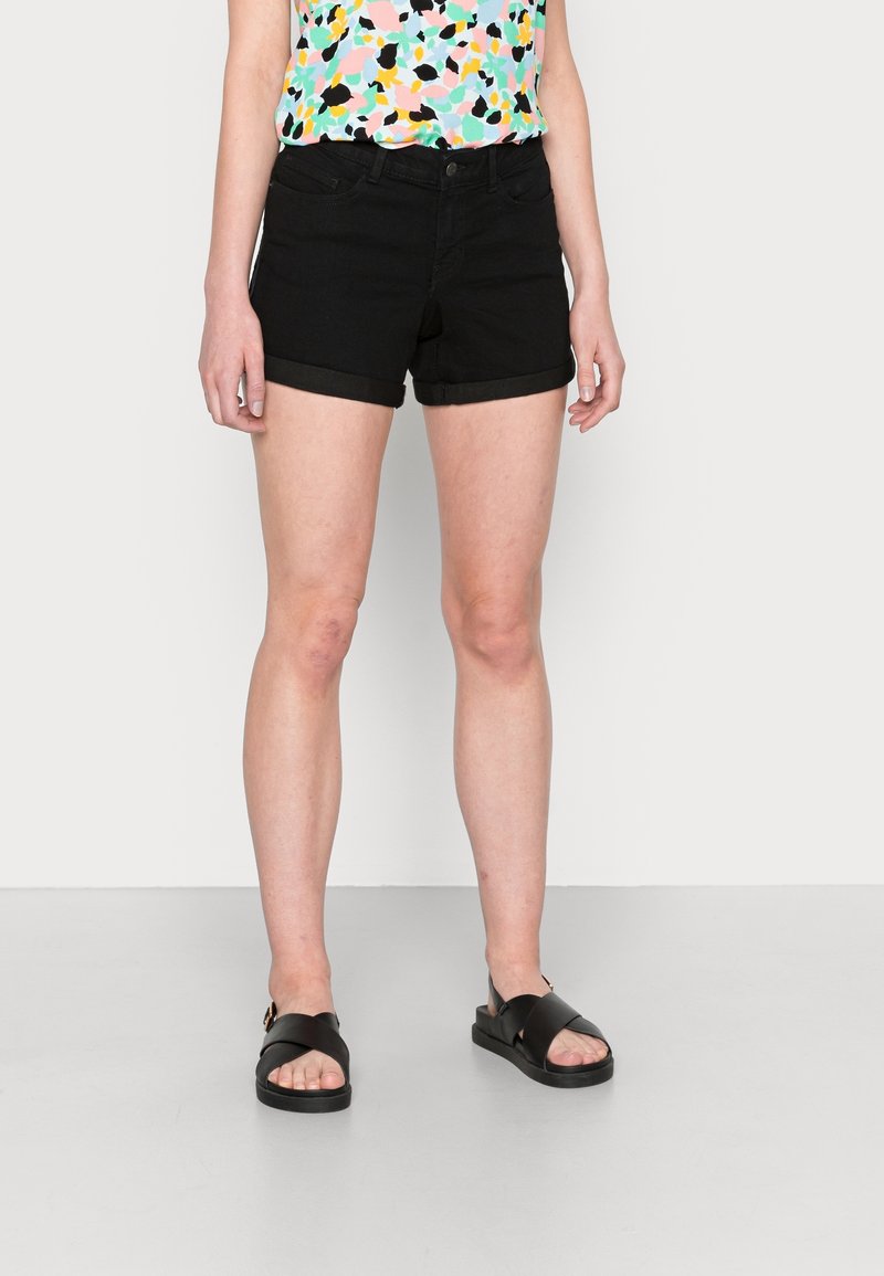 Femme portant un short noir à ourlet roulé, un haut multicolore à motif abstrait et des sandales noires, debout devant un fond uni.